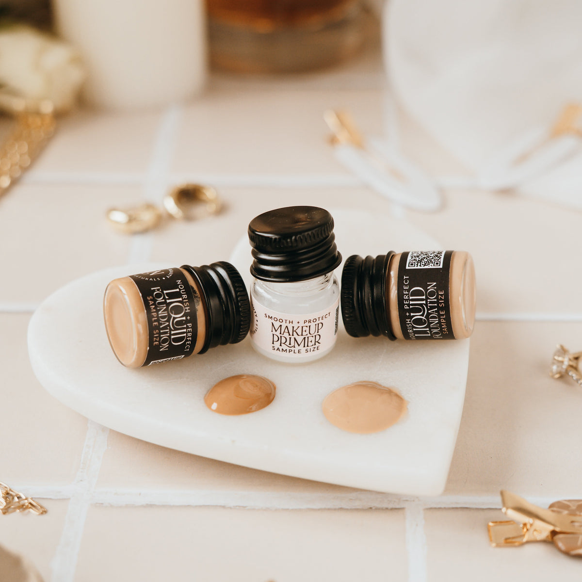 Candid Naturals Smoothing Primer and Foundation bottles with cream swatches on a marble dish, styled with jewelry