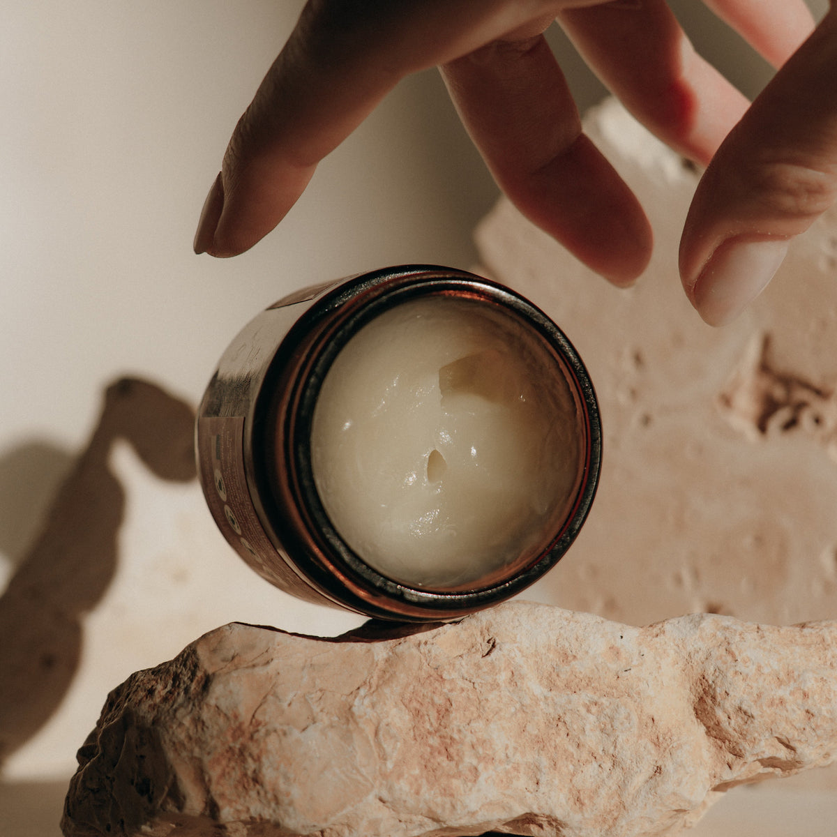 Hand reaching for Candid Naturals Face Balm in an open jar on a pink stone
